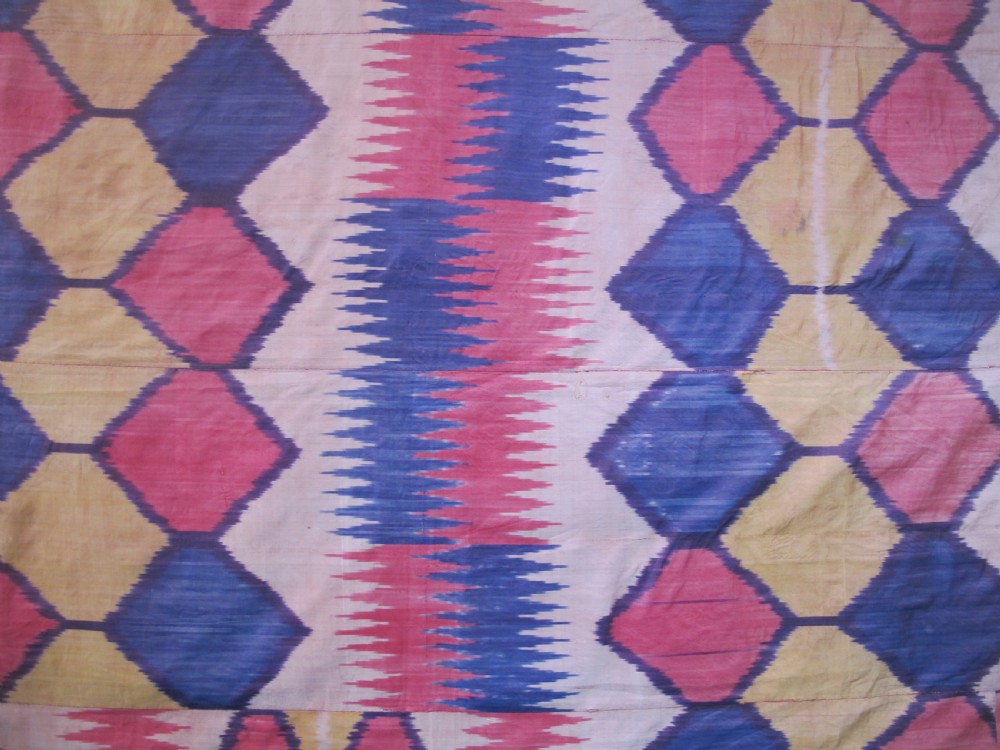 A large handwoven silk ikat textile panel from Uzbekistan featuring geometric diamond motifs and sawtooth patterns in pink, yellow, and purple. View VI