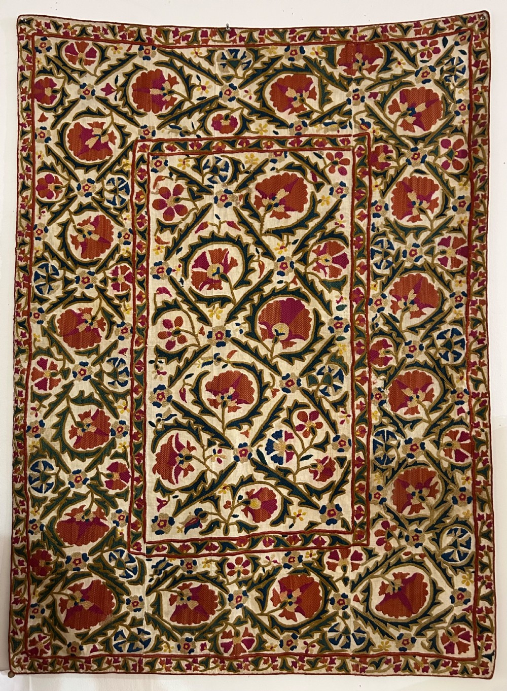 A rectangular silk embroidered suzani panel featuring terracotta red floral blossoms and dark green foliage on a cream woven ground.