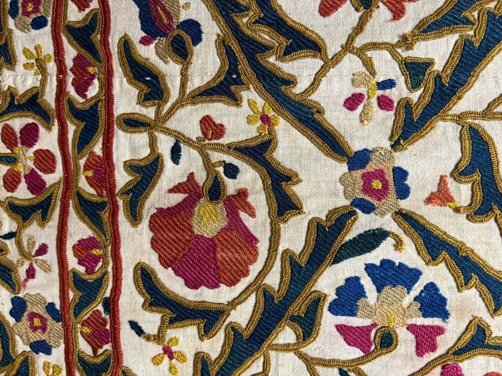A rectangular silk embroidered suzani panel featuring terracotta red floral blossoms and dark green foliage on a cream woven ground. View V