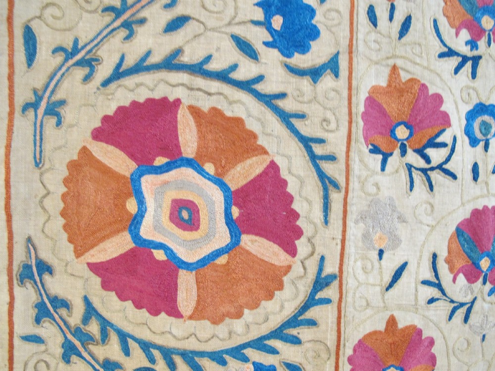 A rectangular 19th-century Bokhara suzani textile featuring hand-embroidered floral rosettes and leaf motifs in red, terracotta, and blue silk threads on a cream cotton field. View VIII