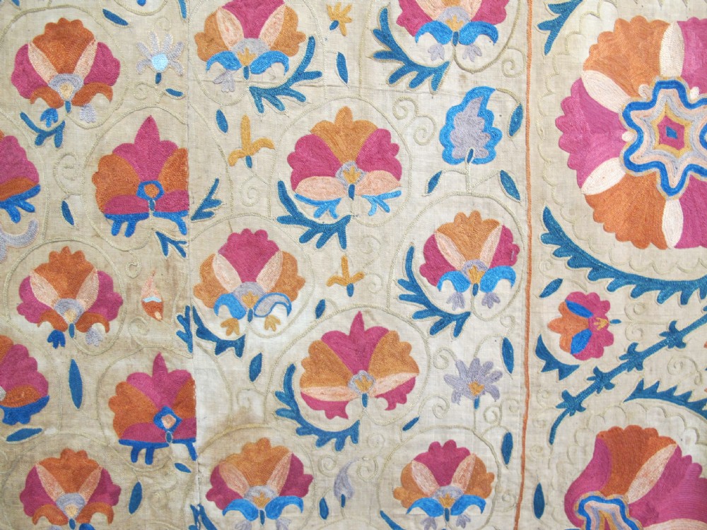 A rectangular 19th-century Bokhara suzani textile featuring hand-embroidered floral rosettes and leaf motifs in red, terracotta, and blue silk threads on a cream cotton field. View X