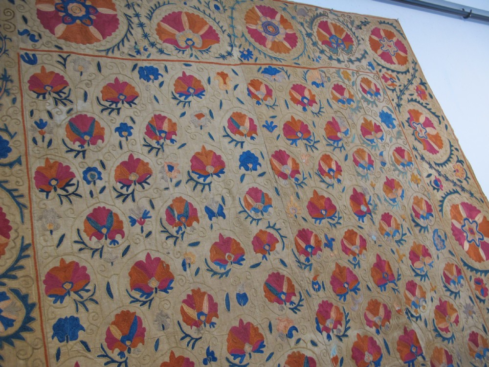 A rectangular 19th-century Bokhara suzani textile featuring hand-embroidered floral rosettes and leaf motifs in red, terracotta, and blue silk threads on a cream cotton field. View XII