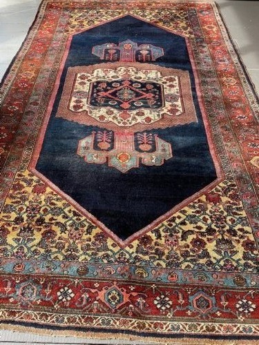 An antique Persian Kurdish wool carpet featuring a deep navy blue central field with a geometric medallion, framed by patterned spandrels and multi-tiered terracotta borders. View V