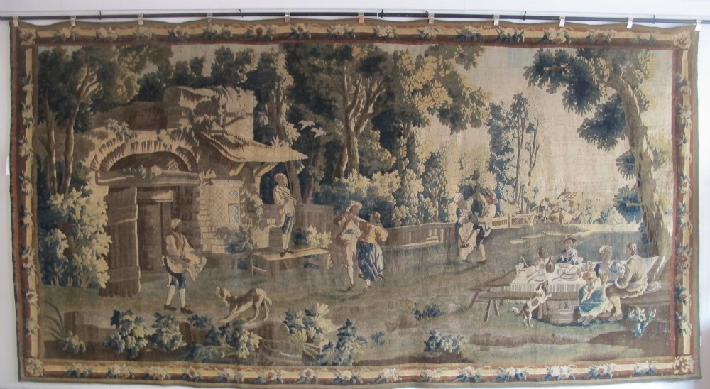 A large late 18th-century French woven tapestry portraying a pastoral scene with dancing figures, a stone building, and leafy trees enclosed within a decorative floral border.