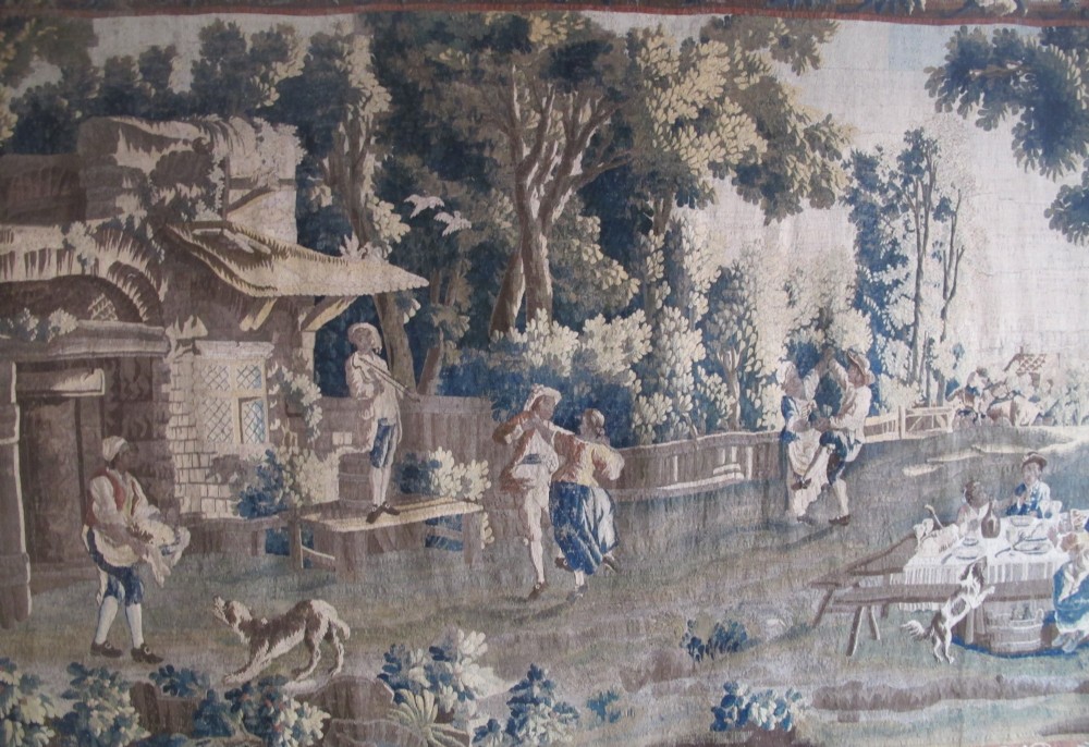 A large late 18th-century French woven tapestry portraying a pastoral scene with dancing figures, a stone building, and leafy trees enclosed within a decorative floral border. View II