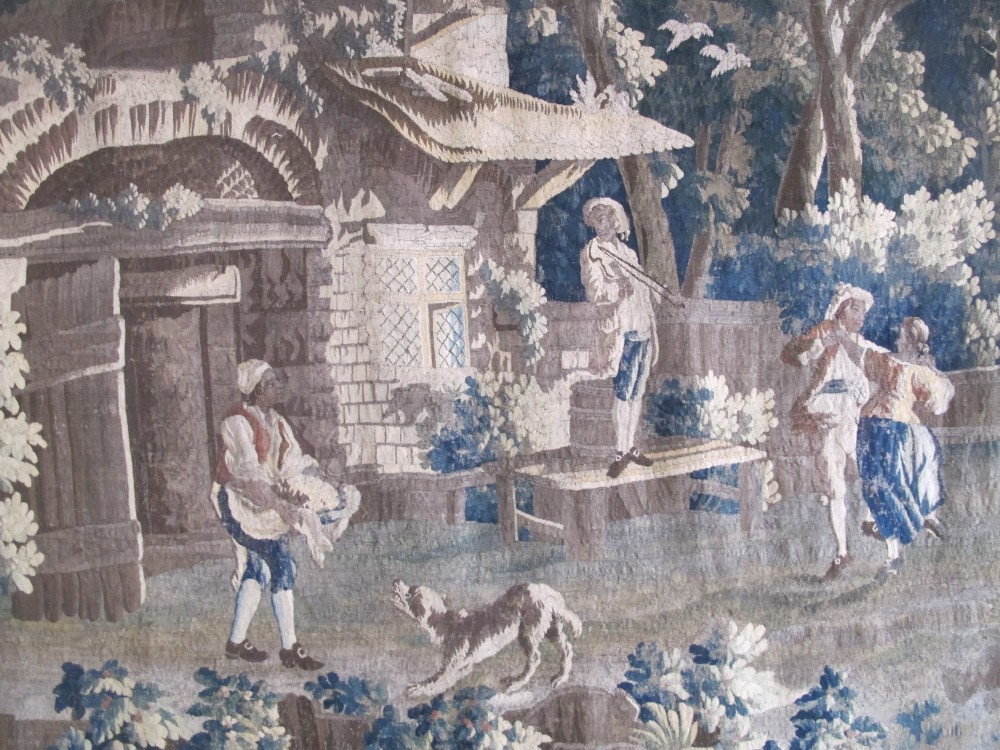 A large late 18th-century French woven tapestry portraying a pastoral scene with dancing figures, a stone building, and leafy trees enclosed within a decorative floral border. View III