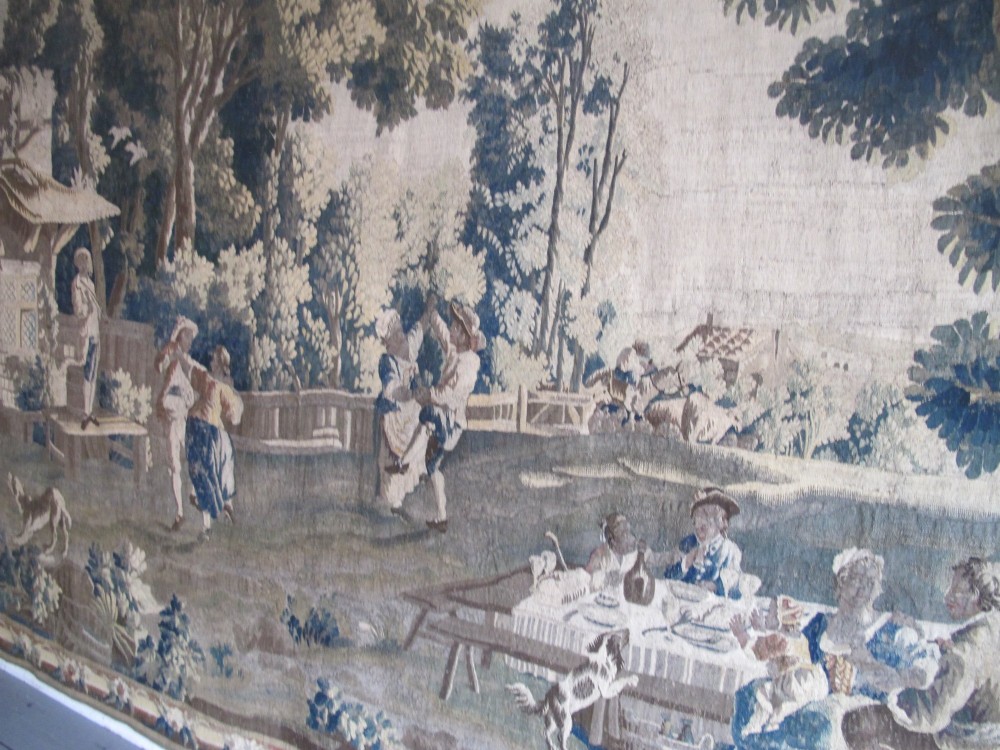 A large late 18th-century French woven tapestry portraying a pastoral scene with dancing figures, a stone building, and leafy trees enclosed within a decorative floral border. View XV