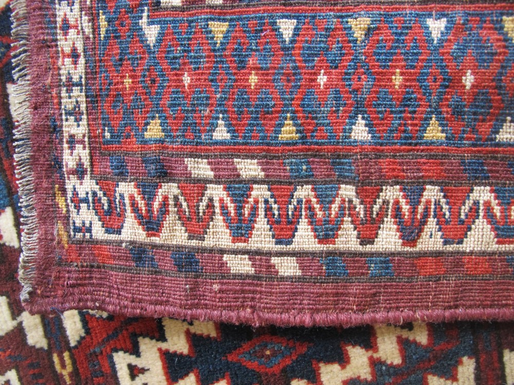 A pentagonal hand-woven wool textile featuring a red and cream geometric diamond lattice pattern with dark navy blue accents. View IV