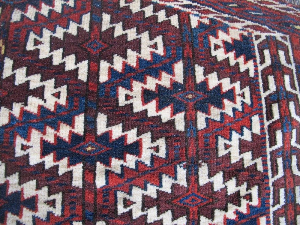 A pentagonal hand-woven wool textile featuring a red and cream geometric diamond lattice pattern with dark navy blue accents. View VI