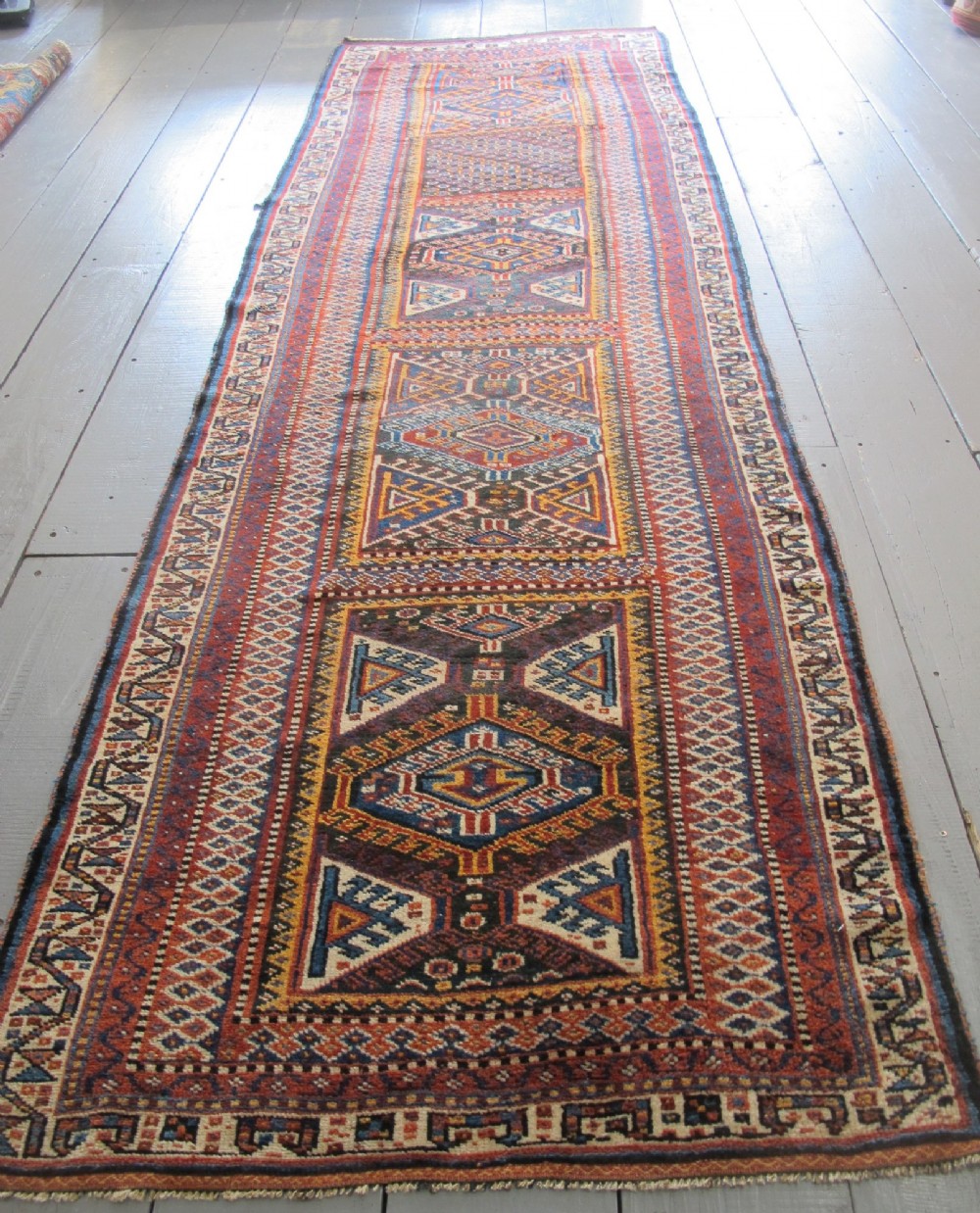 An antique late 19th-century Persian tribal runner rug with repeating geometric diamond medallions and layered borders in terracotta red, indigo blue, and ochre yellow.