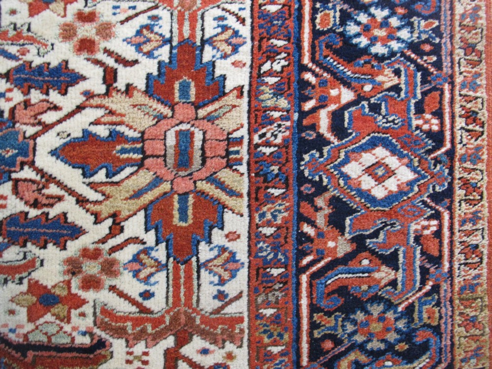 A large hand-knotted Persian Heriz rug featuring a central geometric medallion on an ivory background with terracotta and blue borders. View X