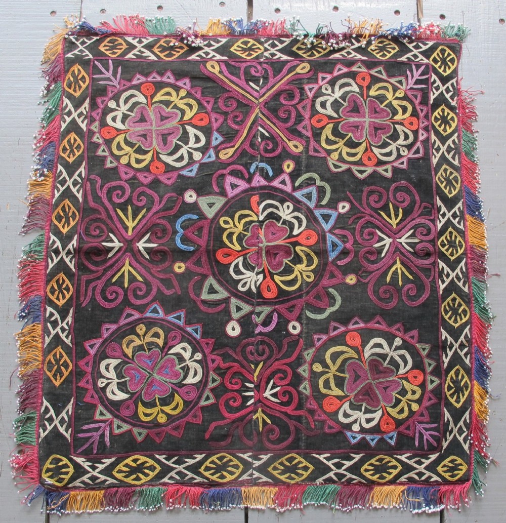 A square dark fabric mirror cover featuring intricate multicoloured silk chain stitch embroidery in circular solar motifs surrounded by a beaded fringe border.
