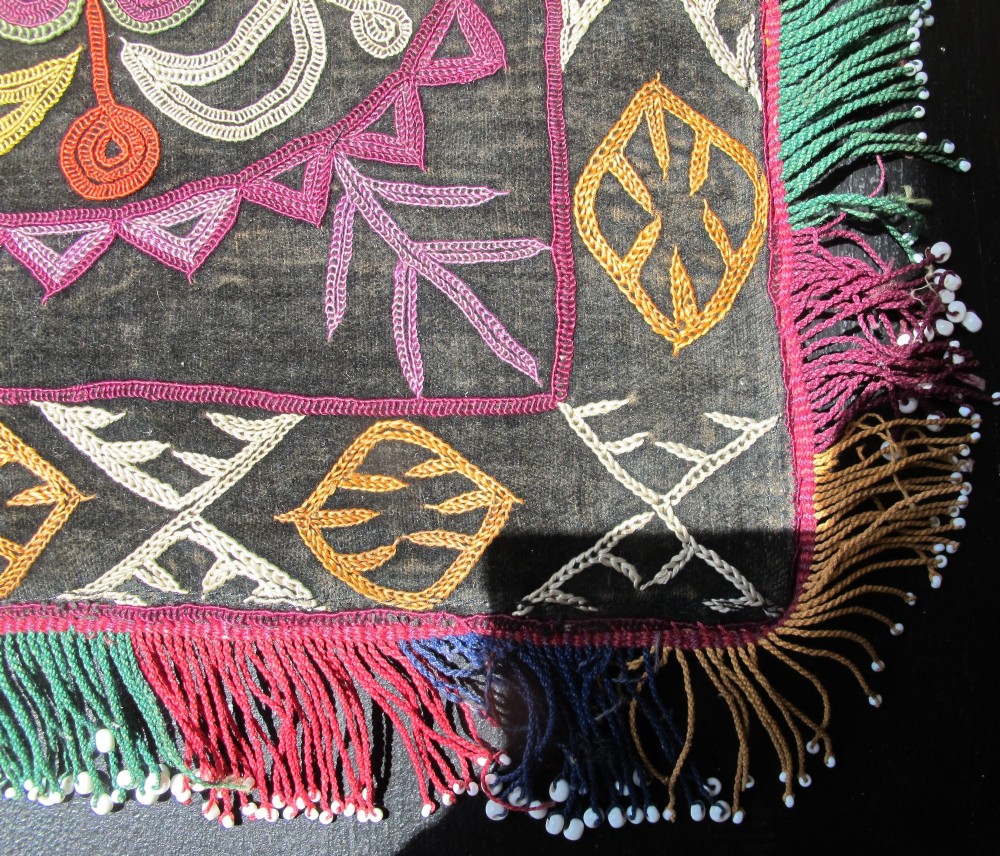 A square dark fabric mirror cover featuring intricate multicoloured silk chain stitch embroidery in circular solar motifs surrounded by a beaded fringe border. View IX