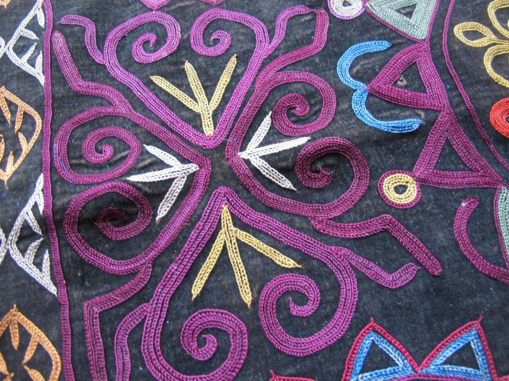 A square dark fabric mirror cover featuring intricate multicoloured silk chain stitch embroidery in circular solar motifs surrounded by a beaded fringe border. View X