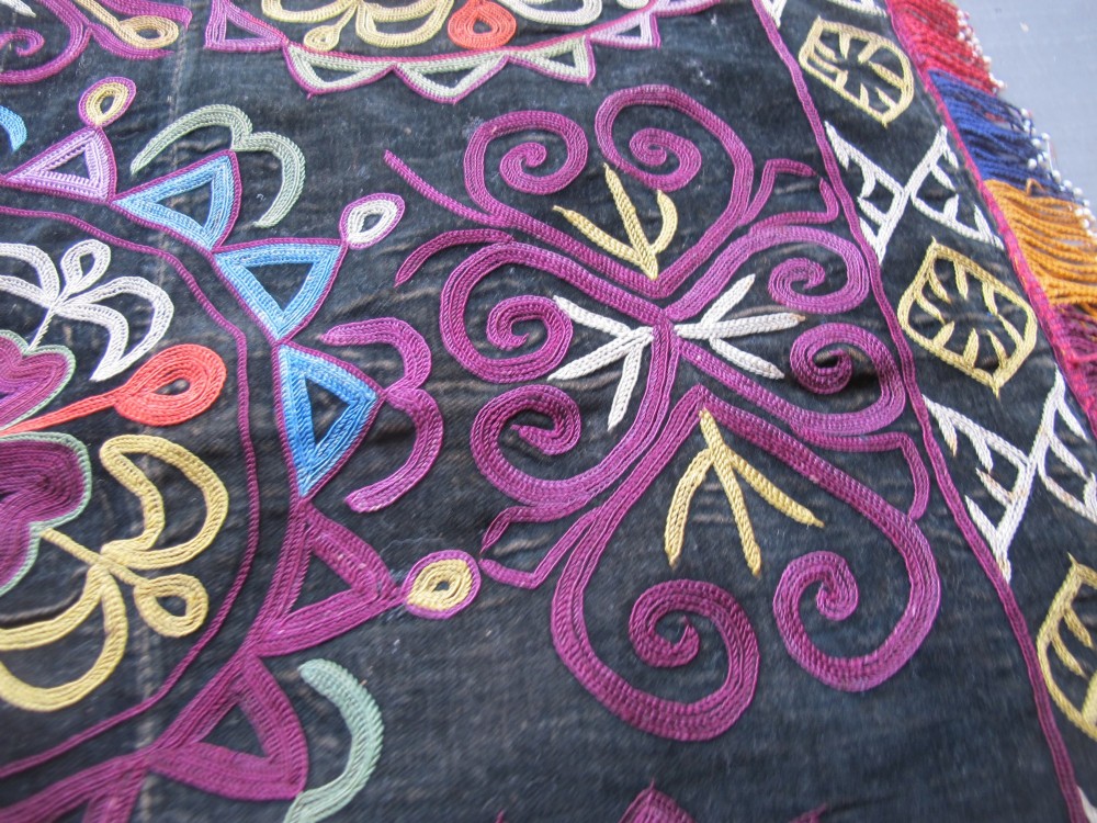 A square dark fabric mirror cover featuring intricate multicoloured silk chain stitch embroidery in circular solar motifs surrounded by a beaded fringe border. View XII