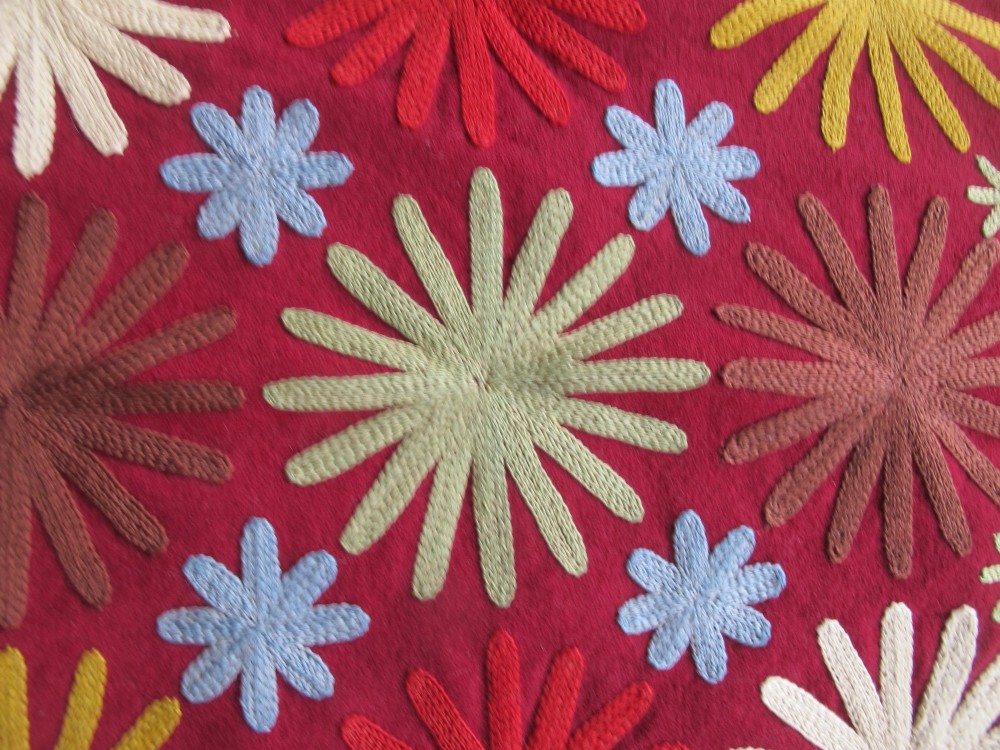 A square red embroidered Lakai textile featuring stylised sunburst and spiral motifs framed by a dark fringe. View V