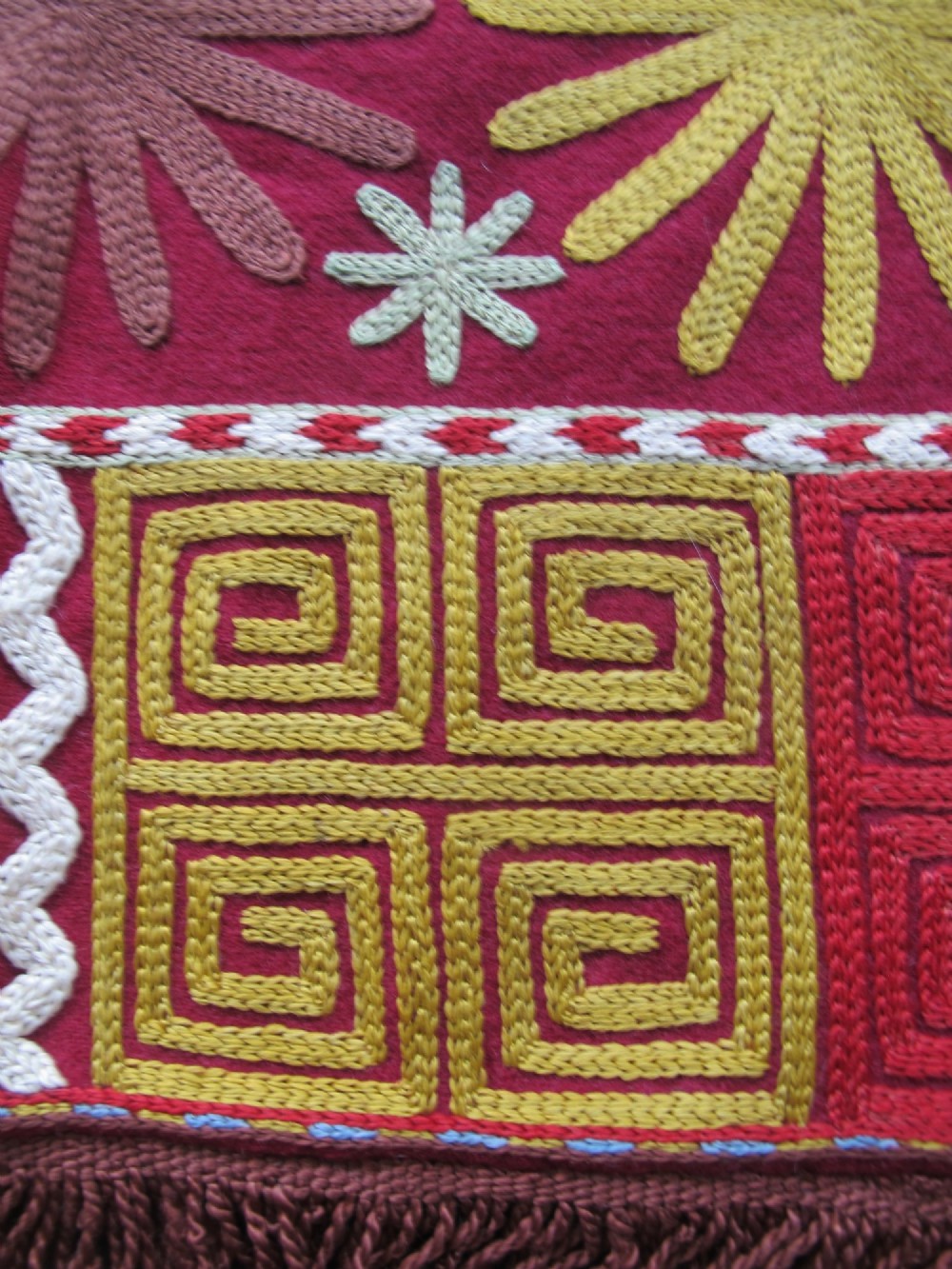 A square red embroidered Lakai textile featuring stylised sunburst and spiral motifs framed by a dark fringe. View VI