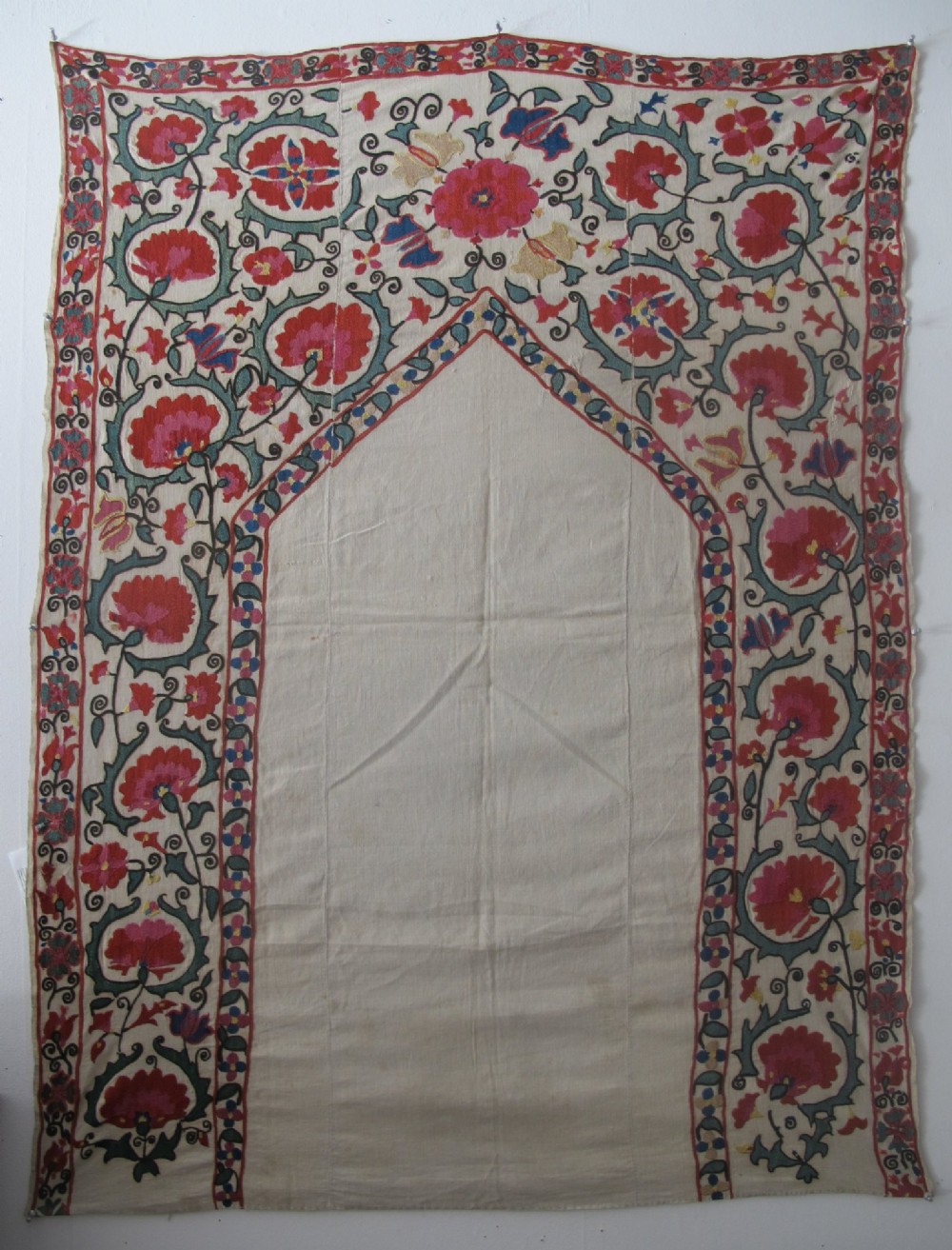 An antique cream cotton suzani textile embroidered with an architectural arch and red floral vines with teal leaves.