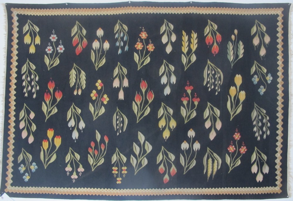 A rectangular black flatwoven wool tapestry featuring repeating stylised colourful botanical flowers and a geometric zigzag border.