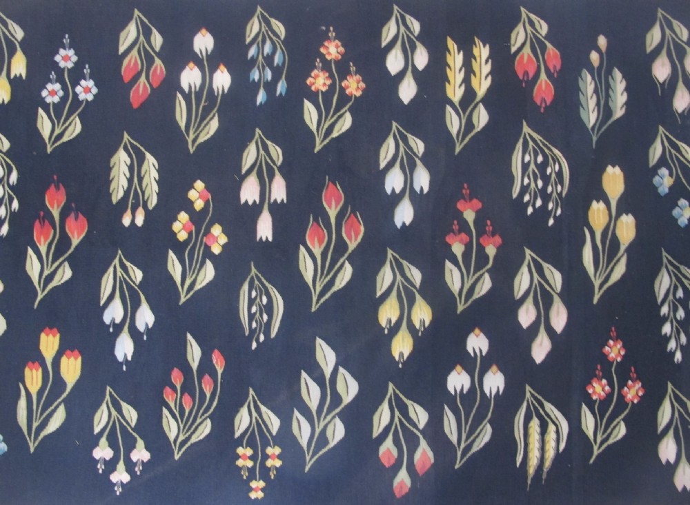 A rectangular black flatwoven wool tapestry featuring repeating stylised colourful botanical flowers and a geometric zigzag border. View II