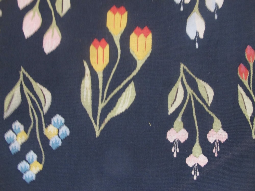 A rectangular black flatwoven wool tapestry featuring repeating stylised colourful botanical flowers and a geometric zigzag border. View III