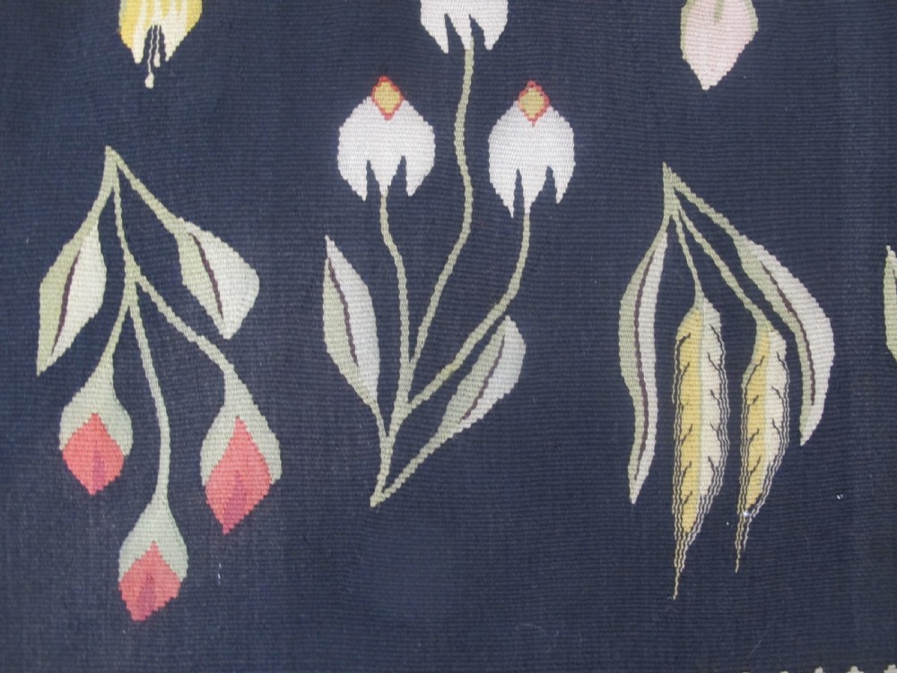 A rectangular black flatwoven wool tapestry featuring repeating stylised colourful botanical flowers and a geometric zigzag border. View IV