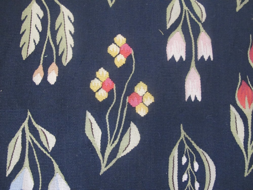 A rectangular black flatwoven wool tapestry featuring repeating stylised colourful botanical flowers and a geometric zigzag border. View V