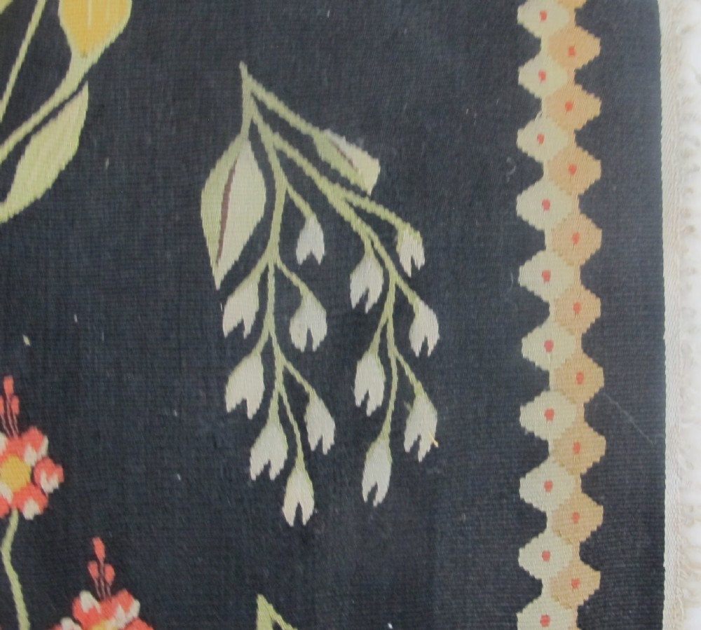 A rectangular black flatwoven wool tapestry featuring repeating stylised colourful botanical flowers and a geometric zigzag border. View VI