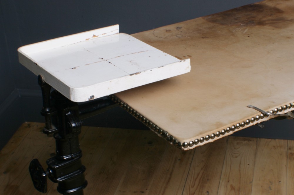 A Victorian cast iron adjustable overbed table featuring a black pedestal stand with castors, a tilting wooden top edged with studding, metal page clips, and a small side tray. View III