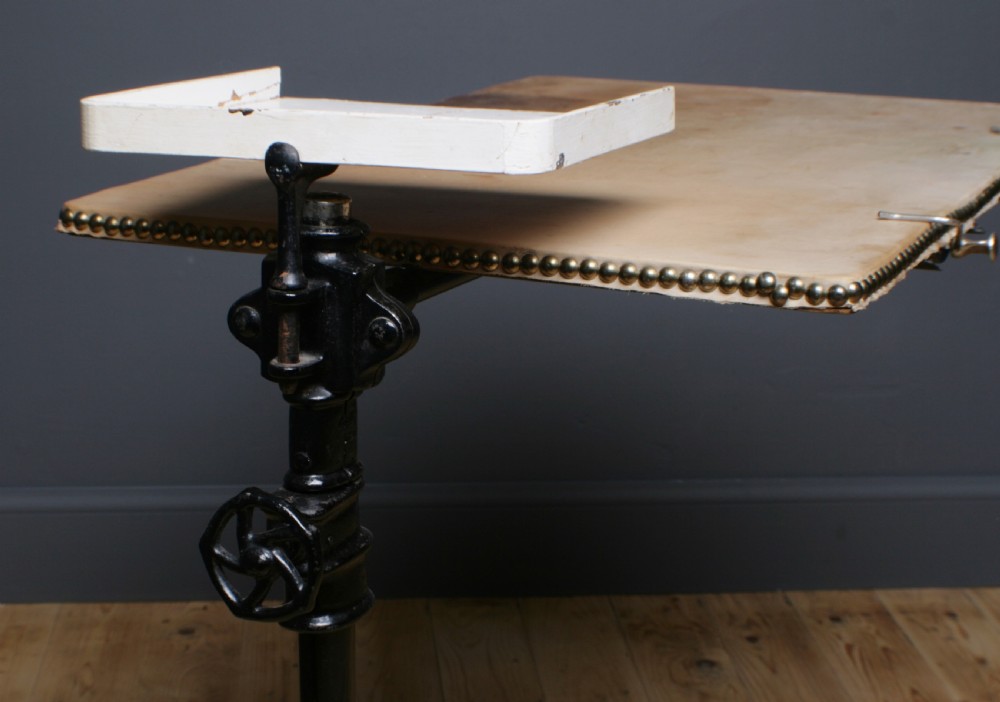 A Victorian cast iron adjustable overbed table featuring a black pedestal stand with castors, a tilting wooden top edged with studding, metal page clips, and a small side tray. View V