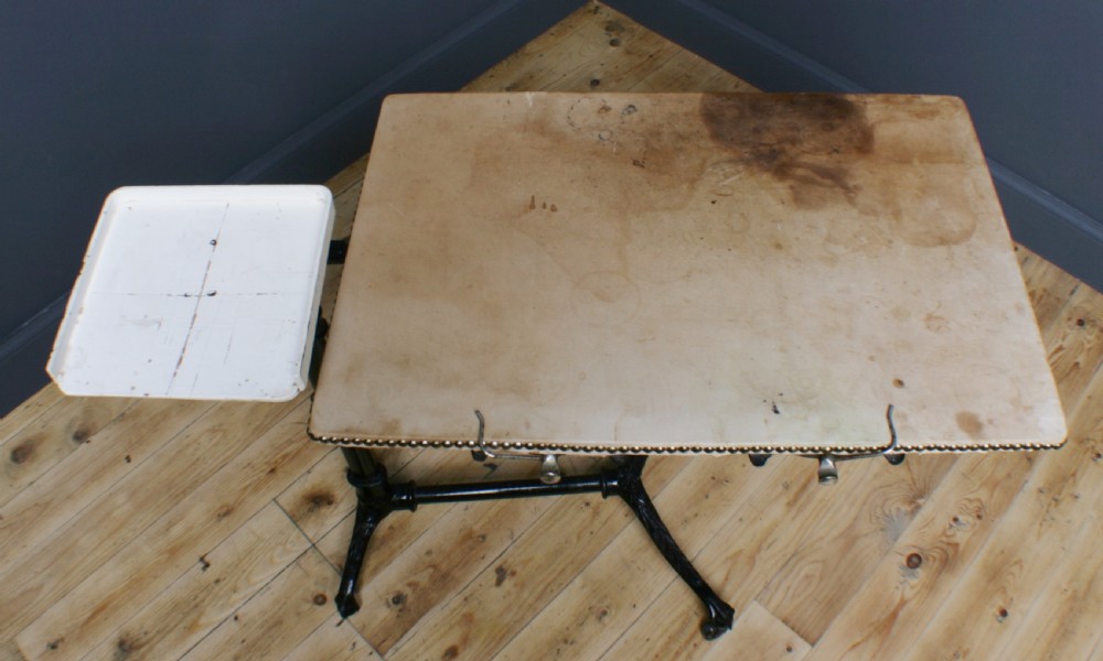 A Victorian cast iron adjustable overbed table featuring a black pedestal stand with castors, a tilting wooden top edged with studding, metal page clips, and a small side tray. View VI