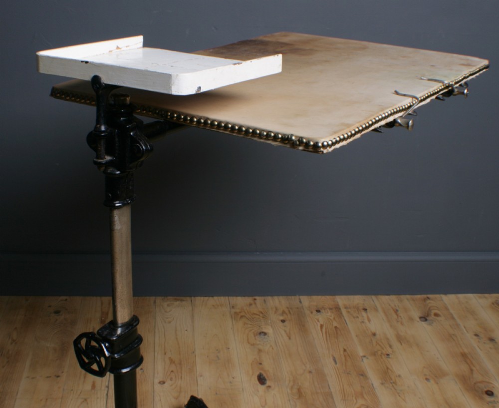 A Victorian cast iron adjustable overbed table featuring a black pedestal stand with castors, a tilting wooden top edged with studding, metal page clips, and a small side tray. View IX