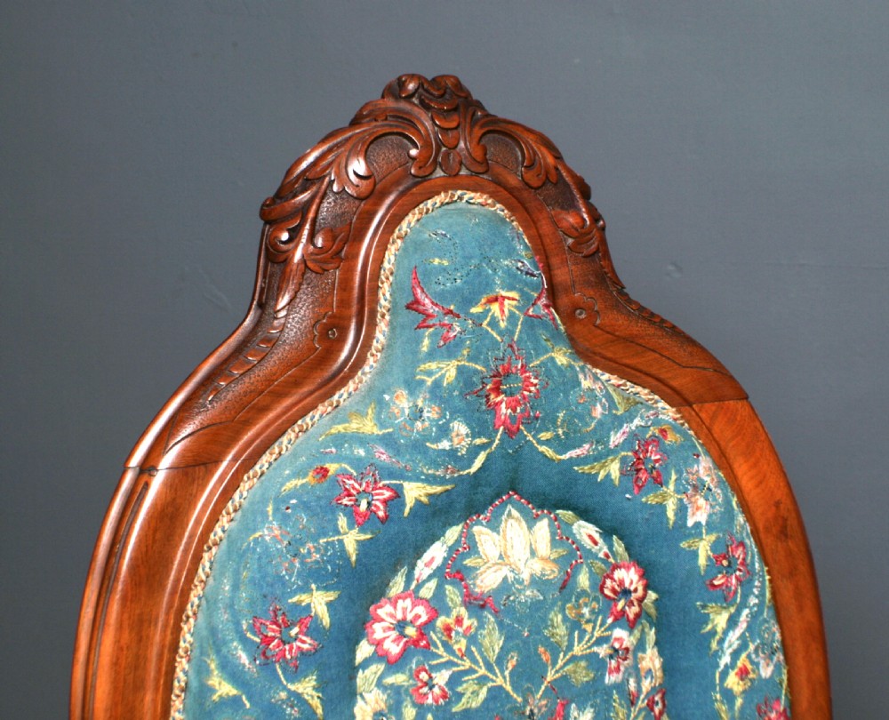 An antique Victorian mahogany nursing chair featuring a carved top crest, blue floral needlepoint upholstery on the back and seat, and turned front legs on ceramic castors. View III