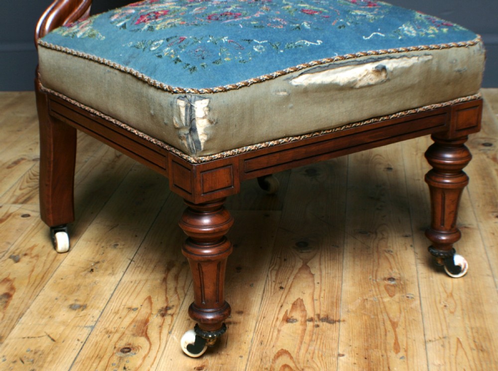 An antique Victorian mahogany nursing chair featuring a carved top crest, blue floral needlepoint upholstery on the back and seat, and turned front legs on ceramic castors. View VI