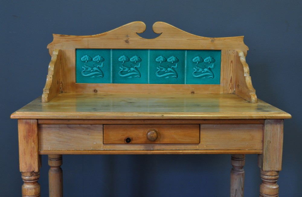 An antique Victorian pine washstand featuring a raised gallery splashback set with four green embossed tiles, a single frieze drawer, and turned legs. View V