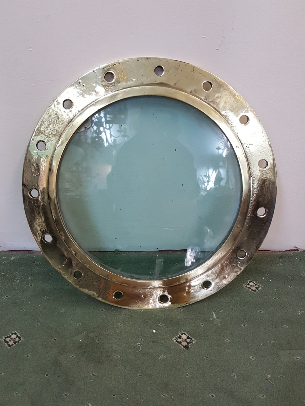 A circular Victorian solid brass ocean liner porthole featuring a central clear glass pane and multiple bolt holes around the wide outer flange.
