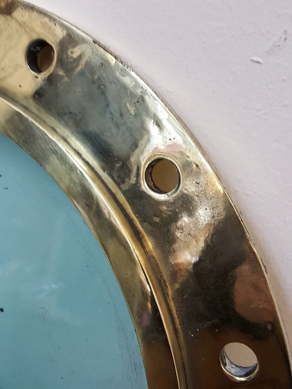 A circular Victorian solid brass ocean liner porthole featuring a central clear glass pane and multiple bolt holes around the wide outer flange. View II