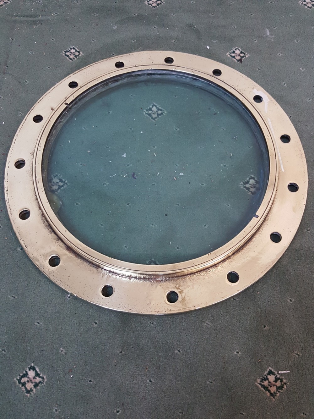 A circular Victorian solid brass ocean liner porthole featuring a central clear glass pane and multiple bolt holes around the wide outer flange. View III