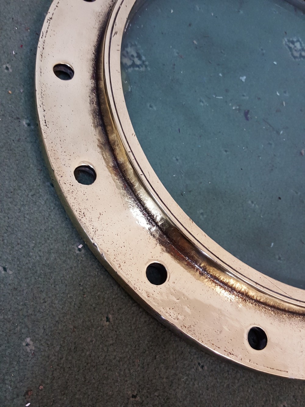 A circular Victorian solid brass ocean liner porthole featuring a central clear glass pane and multiple bolt holes around the wide outer flange. View IV