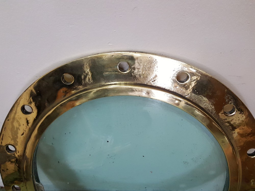 A circular Victorian solid brass ocean liner porthole featuring a central clear glass pane and multiple bolt holes around the wide outer flange. View VI