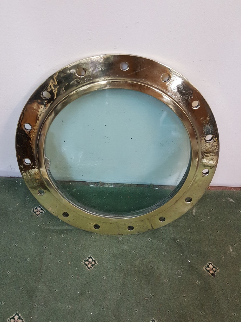 A circular Victorian solid brass ocean liner porthole featuring a central clear glass pane and multiple bolt holes around the wide outer flange. View VII