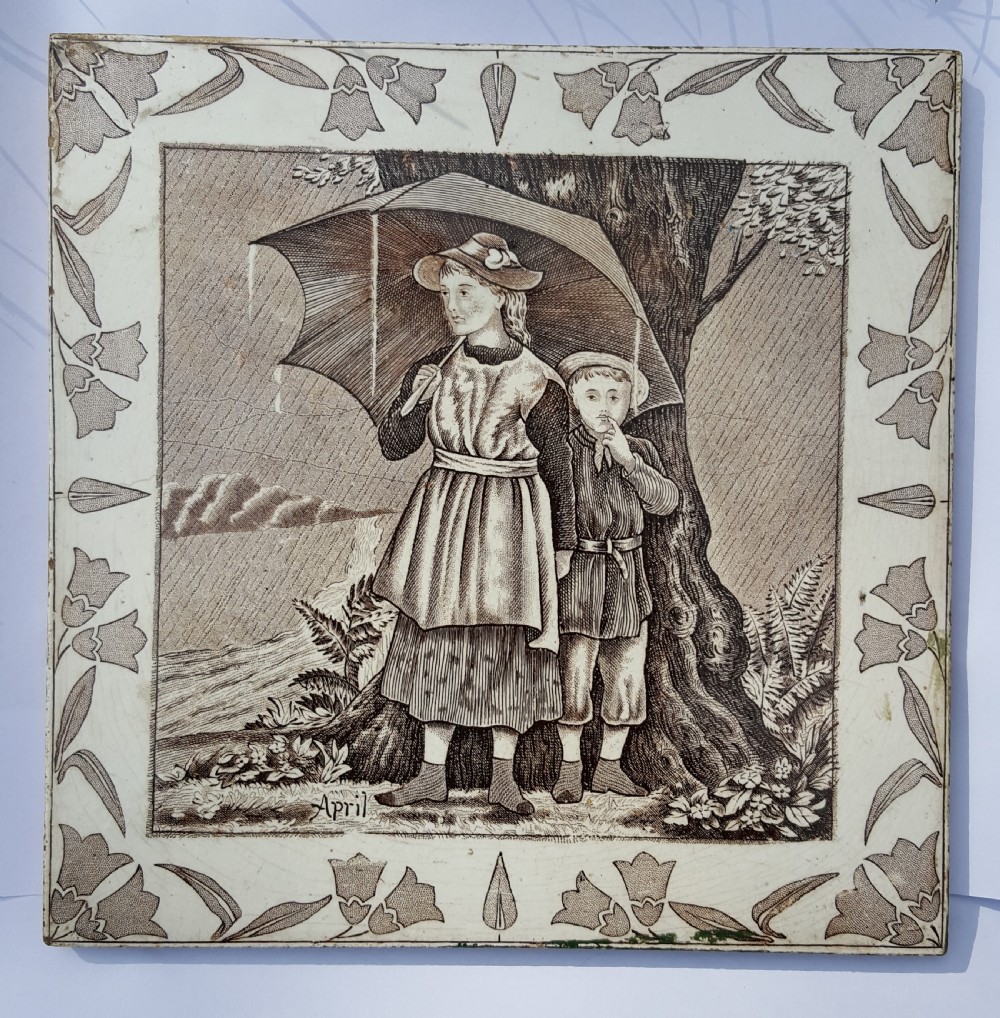 A square Victorian ceramic tile featuring a sepia transfer print titled April, depicting two children sheltering under an umbrella in the rain framed by a botanical floral border.