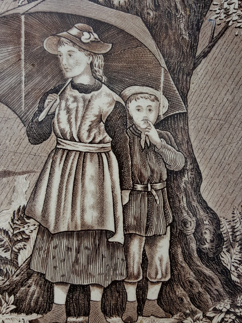 A square Victorian ceramic tile featuring a sepia transfer print titled April, depicting two children sheltering under an umbrella in the rain framed by a botanical floral border. View II