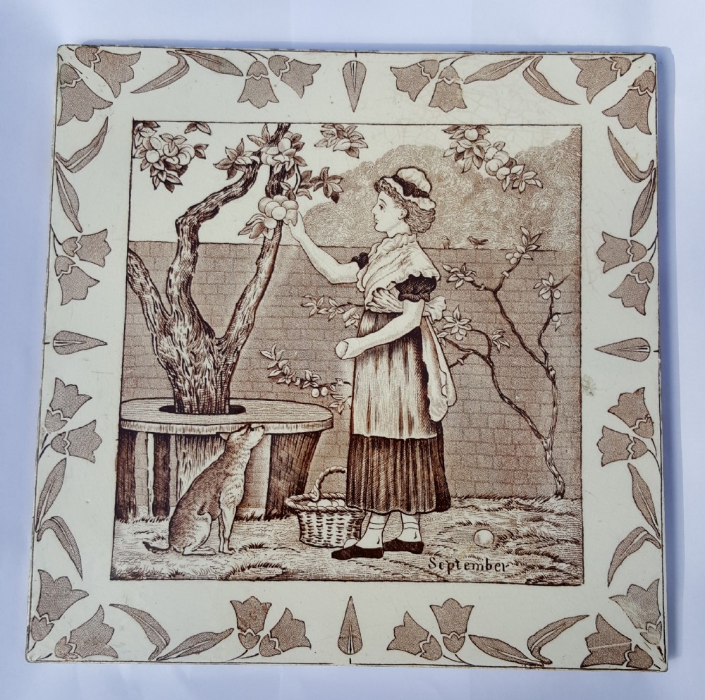 A square Victorian ceramic tile featuring a sepia transfer print of a female figure harvesting fruit under a tree with September written below, surrounded by a foliage border.