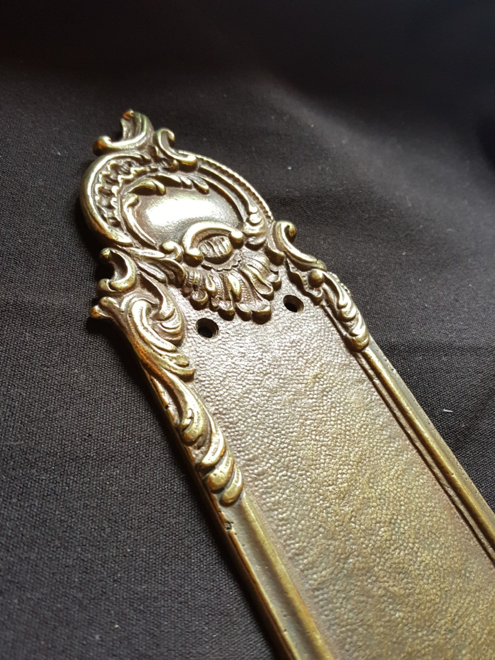 A single cast brass Georgian style finger plate displaying scrollwork and cartouche relief detailing at top and bottom ends. View II