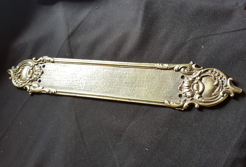 A single cast brass Georgian style finger plate displaying scrollwork and cartouche relief detailing at top and bottom ends. View IV