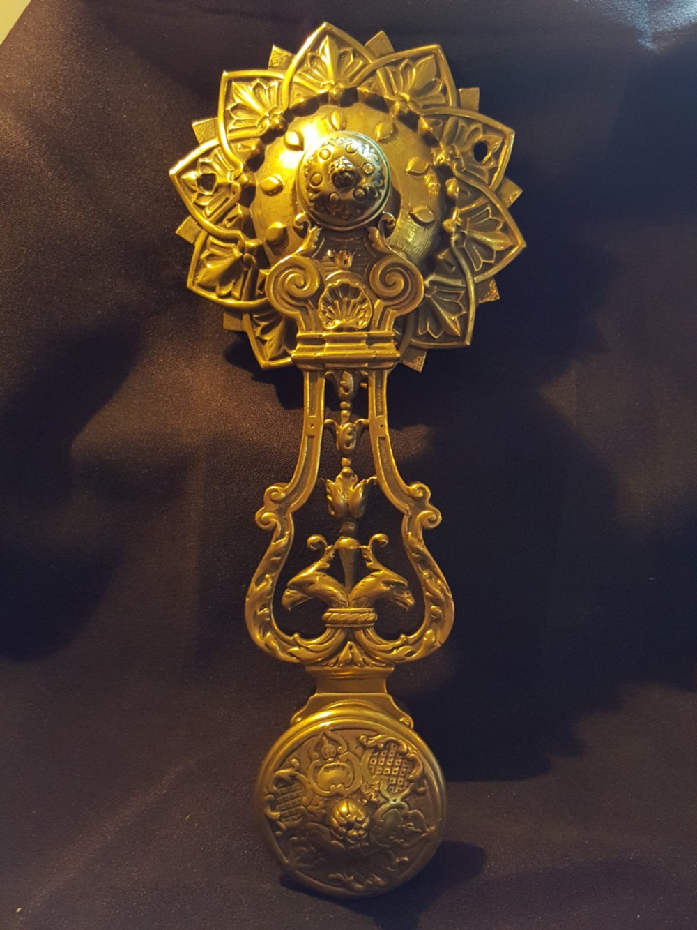 An ornate cast brass mechanical wall-mounted bell ringer featuring a sunburst rosette top, openwork scrollwork with mythical creatures, and a circular decorative pendulum drop.