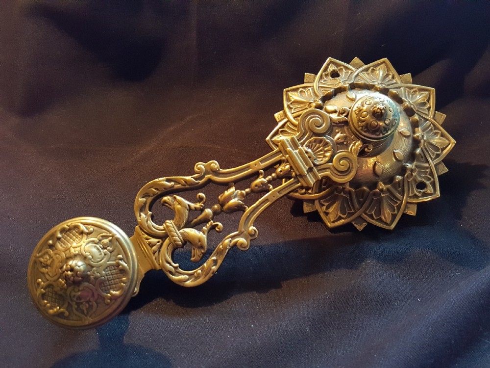 An ornate cast brass mechanical wall-mounted bell ringer featuring a sunburst rosette top, openwork scrollwork with mythical creatures, and a circular decorative pendulum drop. View II