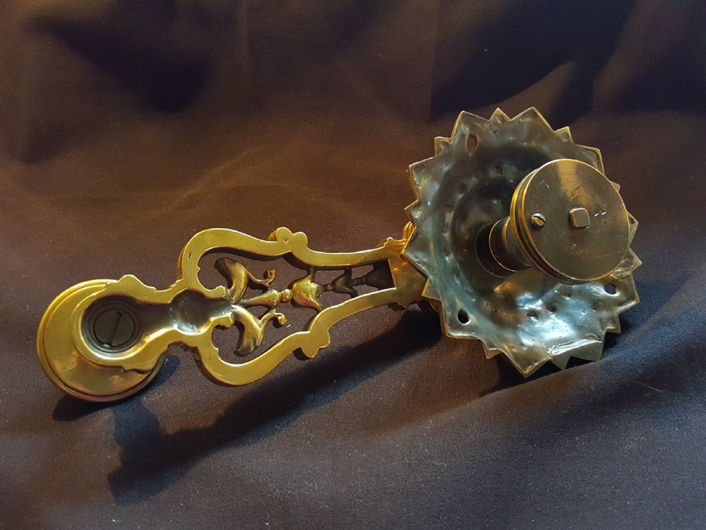 An ornate cast brass mechanical wall-mounted bell ringer featuring a sunburst rosette top, openwork scrollwork with mythical creatures, and a circular decorative pendulum drop. View IV