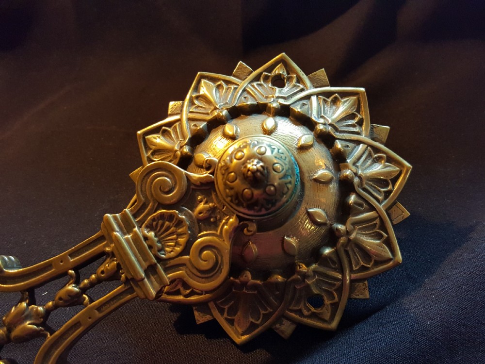 An ornate cast brass mechanical wall-mounted bell ringer featuring a sunburst rosette top, openwork scrollwork with mythical creatures, and a circular decorative pendulum drop. View V